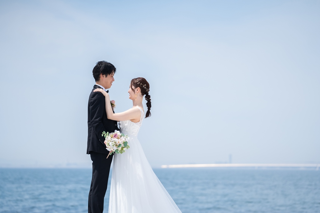 水平線が広がる海を背景に、見つめ合うタキシードの新郎と編み込みヘアの花嫁。爽やかな海辺のウェディングフォト