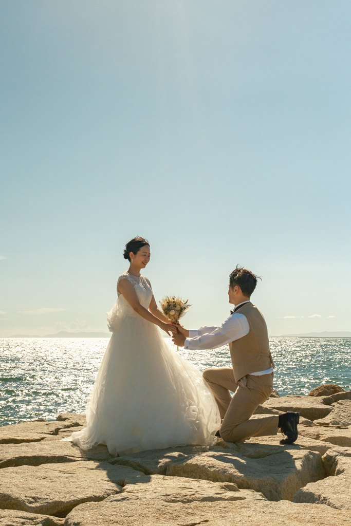 海を背に岩場でひざをつき花嫁の手を取る新郎と、ブーケを持って微笑む花嫁