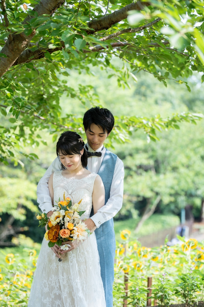 大きな木の木陰で後ろから花嫁を抱きしめる新郎と、オレンジのブーケを胸に抱く花嫁。ひまわりが咲く夏の前撮り