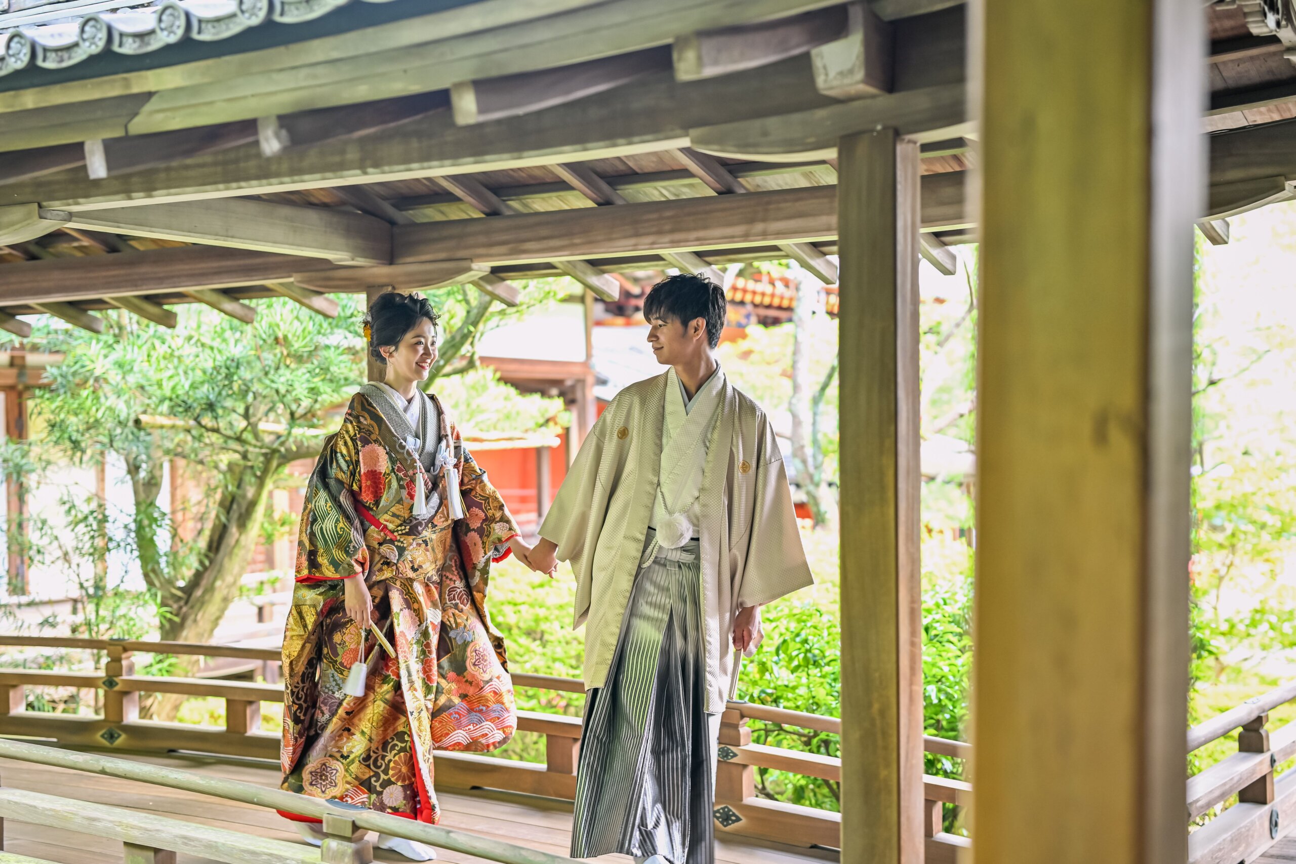 木の柱が連なる寺院の渡り廊下で手を取り合う、色打掛の花嫁とグレーの羽織袴の新郎の和装前撮りの定番ポーズ