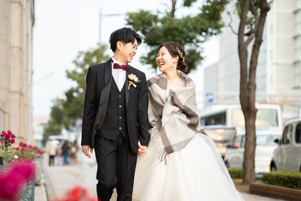 冬の街路樹の下で手をつないで笑い合う、チェックのストールを羽織ったウェディングドレスの花嫁とブラックスーツの新郎の前撮り