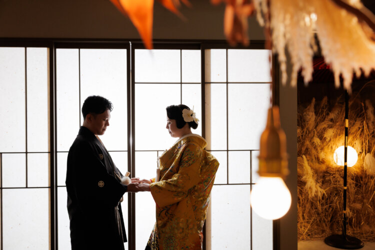 障子窓の前で手を取り合い、向かい合う紋付袴の新郎と色打掛の花嫁。裸電球の灯りがあたたかい和装前撮り