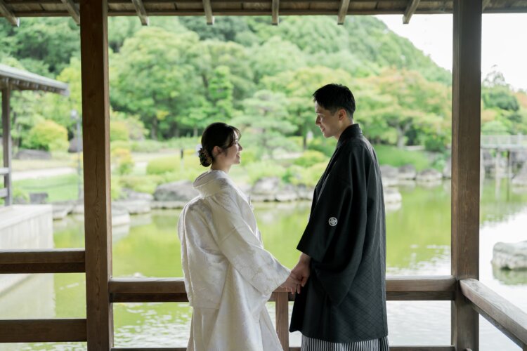 池を望む東屋で手を取り見つめ合う、白無垢の花嫁と紋付袴の新郎。大仙公園日本庭園の新緑に包まれた和装前撮り