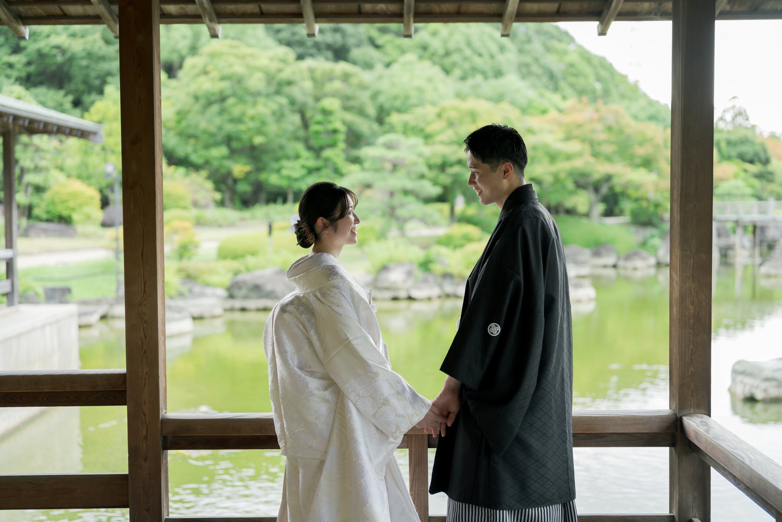 池を望む東屋で手を取り見つめ合う、白無垢の花嫁と紋付袴の新郎。大仙公園日本庭園の新緑に包まれた和装前撮り