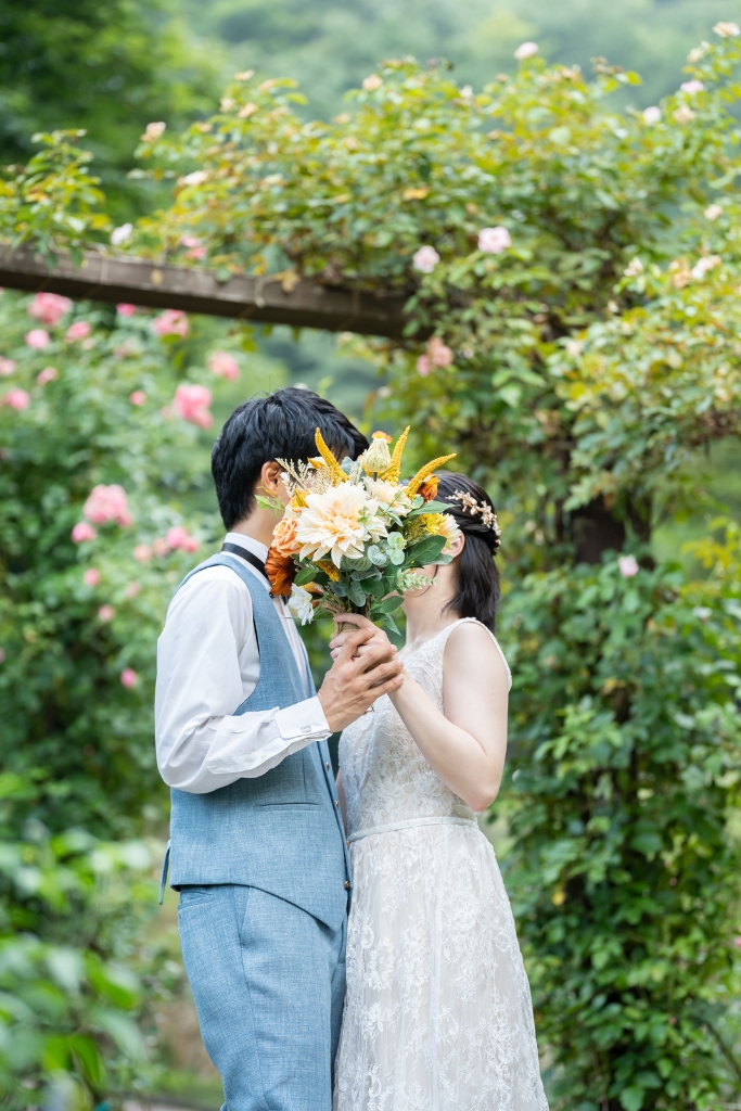 バラのアーチの下でブーケに顔を隠してキスをする新郎新婦。布引ハーブ園の緑に映えるフォトウェディングの人気ポーズ