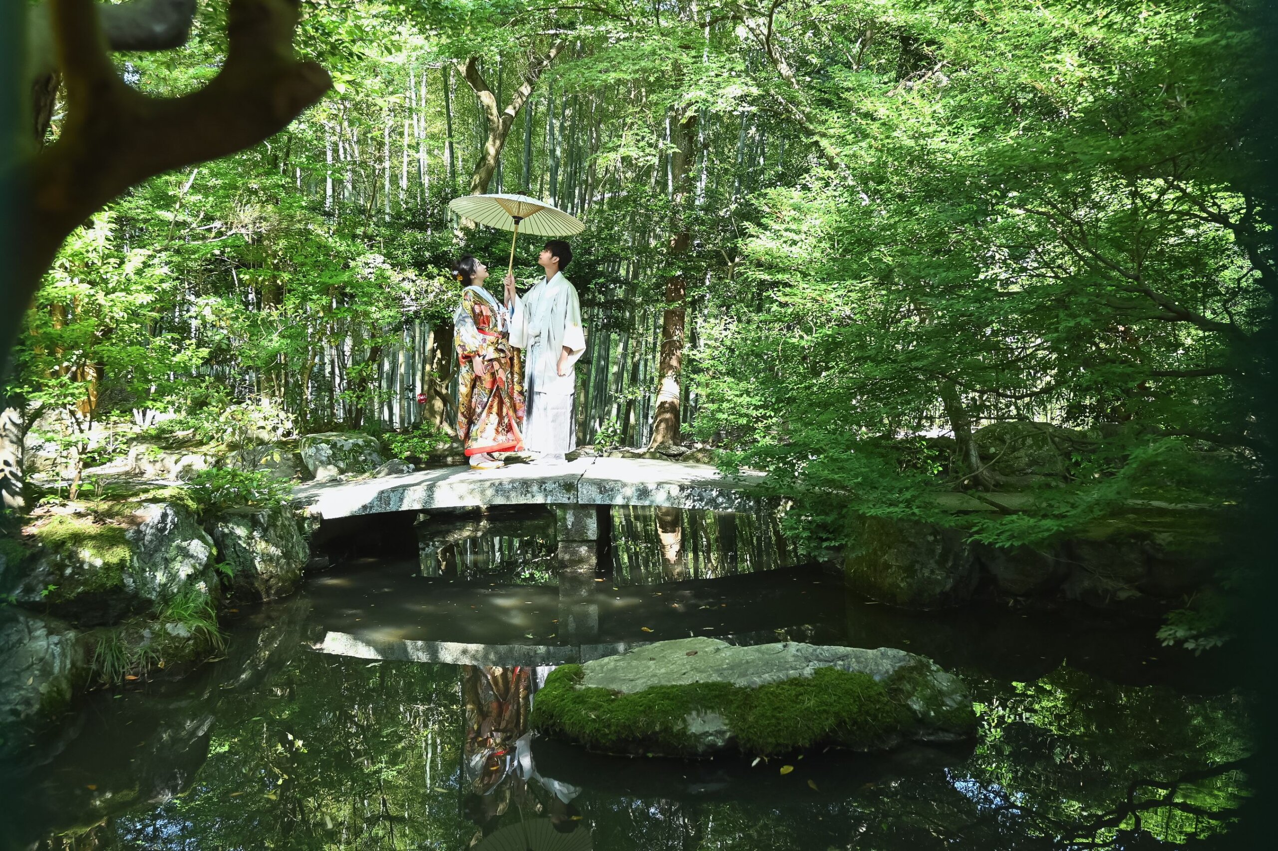 竹林を背にした池の石橋に和傘を差して立つ新郎新婦と、水鏡に映る姿が美しい毘沙門堂の日本庭園での和装前撮り