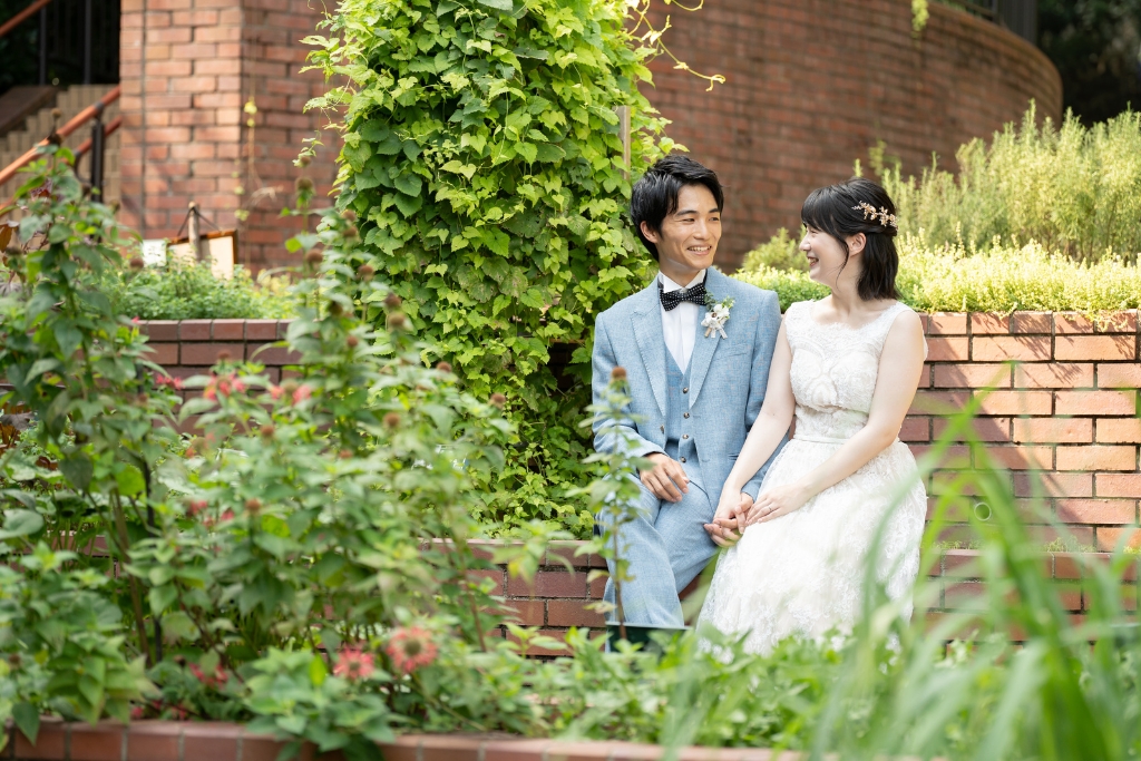 布引ハーブ園のレンガの花壇に並んで腰かけ、緑に囲まれて笑い合うブルーのスーツの新郎とウェディングドレスの花嫁の前撮り