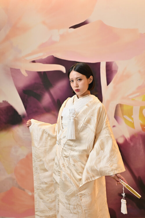 大きな花のアートウォールの前で袖を広げ、扇子を持って立つ金の鶴柄の白無垢の花嫁。色彩豊かな背景が映えるスタジオフォト