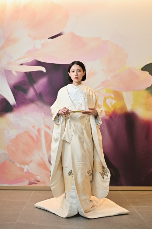 花のアートウォールの前で扇子を持ち、裾を広げて正面に立つ白無垢の花嫁。羽根模様が裾に入った白無垢の全身が分かる結婚写真