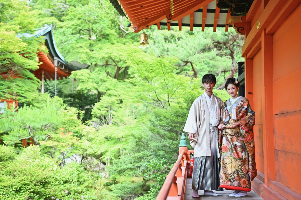 京都の毘沙門堂の朱色の回廊で、紋付袴の新郎と色打掛の花嫁が新緑を背に並ぶ和装の前撮り