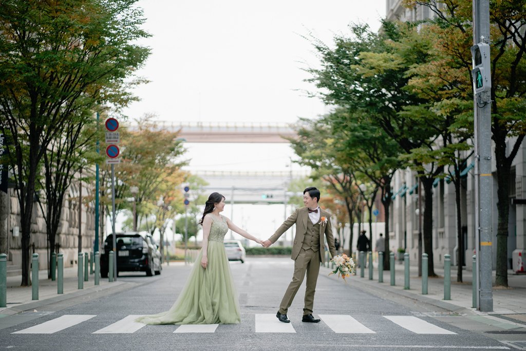 街路樹の並ぶ横断歩道の真ん中で手をつないで見つめ合う、グリーンのカラードレスの花嫁と新郎のロケーションフォト