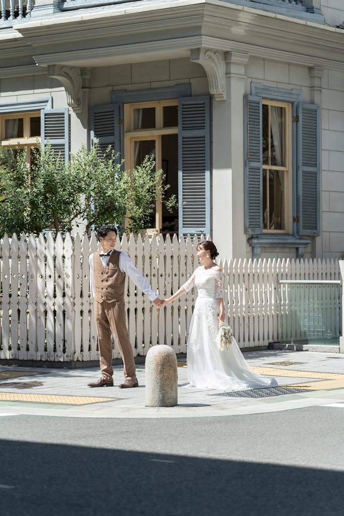 白い柵と青い鎧戸の洋館の前で手をつなぐ、レースのドレスの花嫁とベスト姿の新郎のフォトウェディング