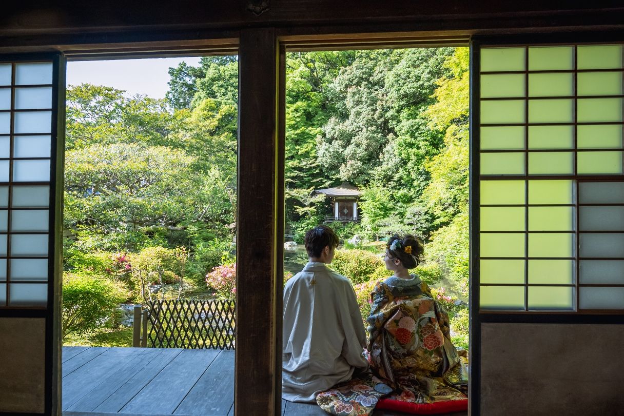 開け放った障子の向こうの庭園を眺めて縁側に座る、色打掛の花嫁と白い羽織袴の新郎の後ろ姿の和装前撮り