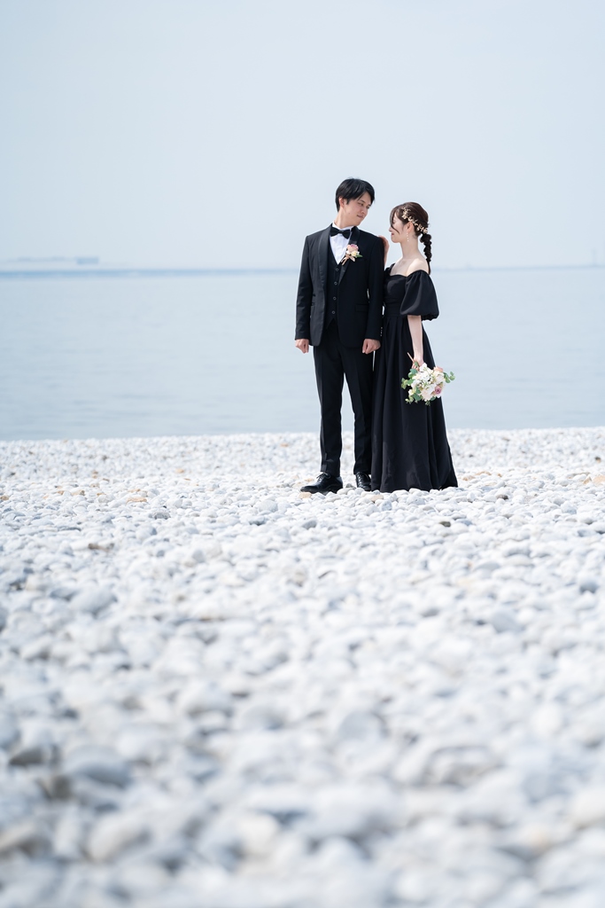 白い小石のビーチで、黒いドレスとタキシードをまとい静かな海を背に見つめ合うふたり