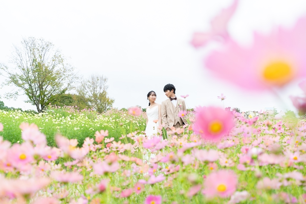 太陽の塔を背景に、見つめ合いながら手を取り合うタキシードの新郎とウェディングドレスの花嫁