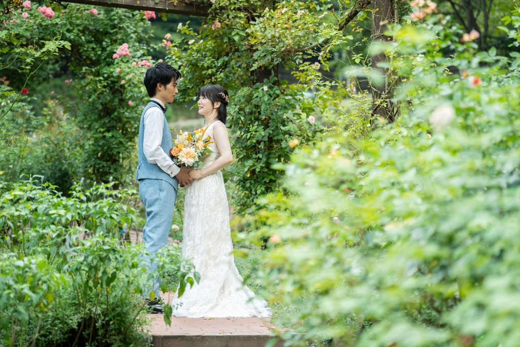 布引ハーブ園のバラの小道で向かい合い、ブーケを手に見つめ合うウェディングドレスの花嫁とベスト姿の新郎のフォトウェディング
