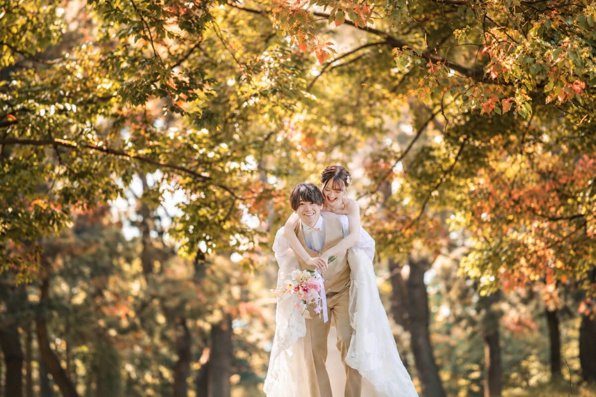 黄色や橙に色づいた並木道で花嫁をおんぶして笑い合う新郎。紅葉フォトウェディング